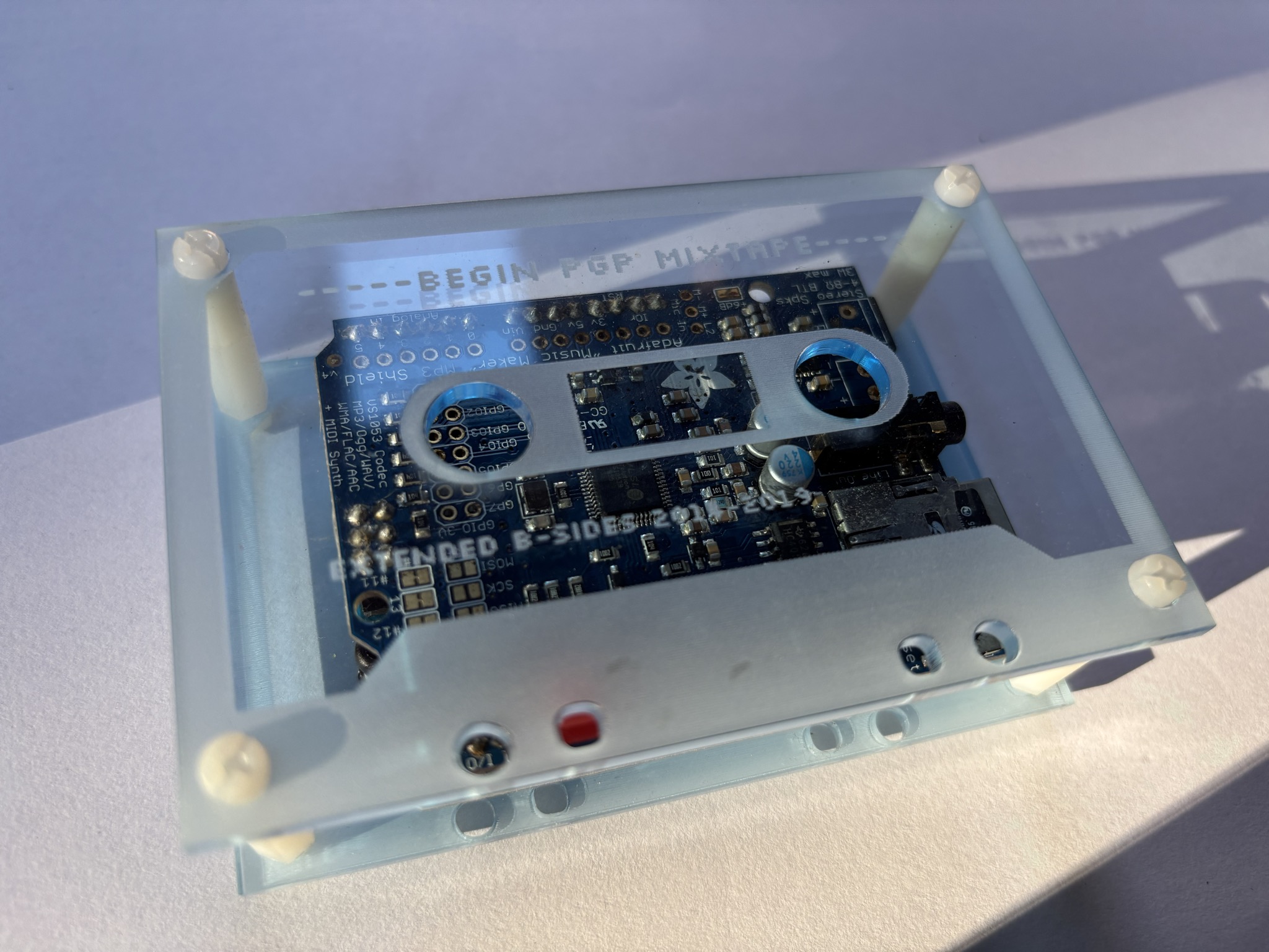 An Arduino microcontroller and music player circuit board in a blue-tinted transparent acrylic enclosure laser-etched to look like a cassette tape.