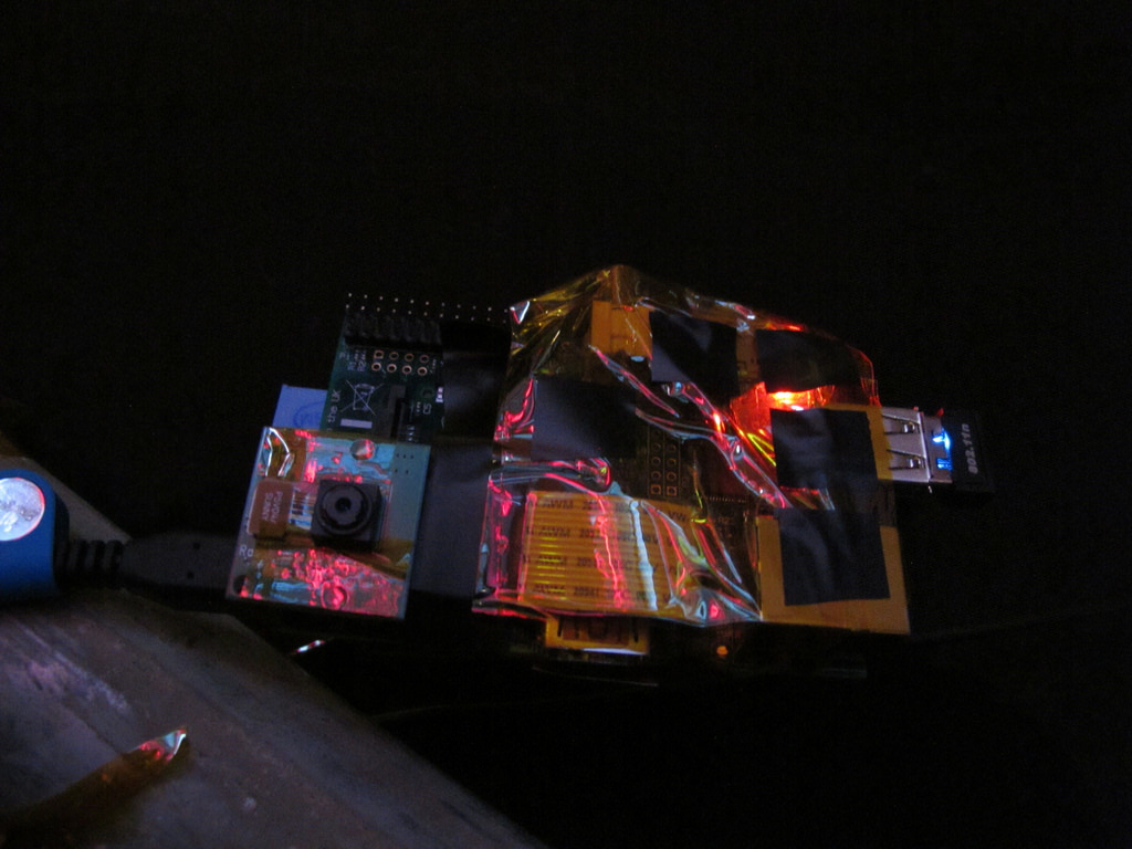 A Raspberry Pi computer and camera with other components wrapped in a cacoon of yellow kapton tape with a 'glider' pattern from Conway's Game of Life made with pieces of electrical tape