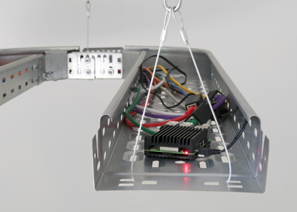 A Raspberry Pi computer in a black metal case with cables coming out the back of it, suspended in an overhead cable raceway similar to those used in data centers.
