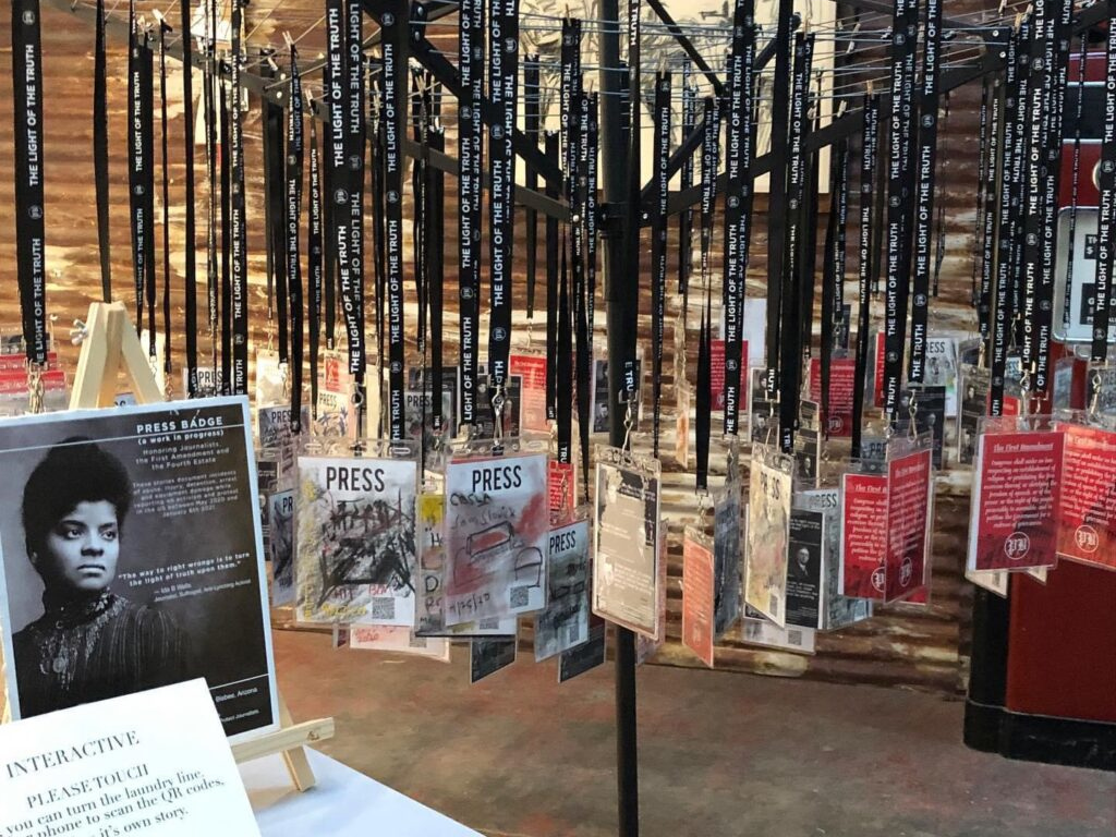 A circular clothes hanger with several lanyards holding informational 'press badges' each featuring a journalist in the U.S. Press Freedom Tracker. A photo of Ida B Wells is on a small table in the foreground.